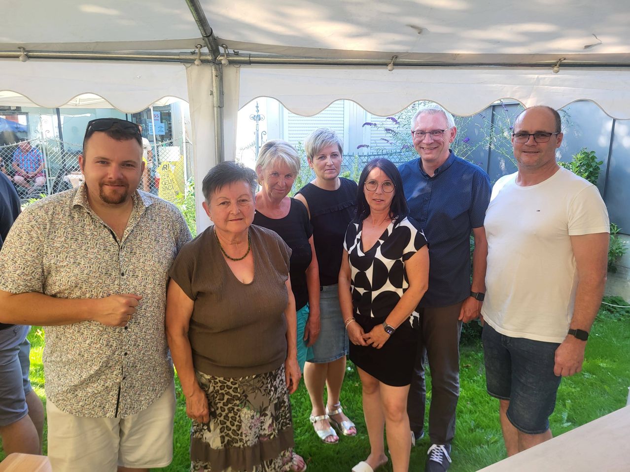 Eine Gruppe von Menschen steht unter einem weißen Zelt in einem Garten. Sie lächeln alle und scheinen für ein Foto zu posieren.