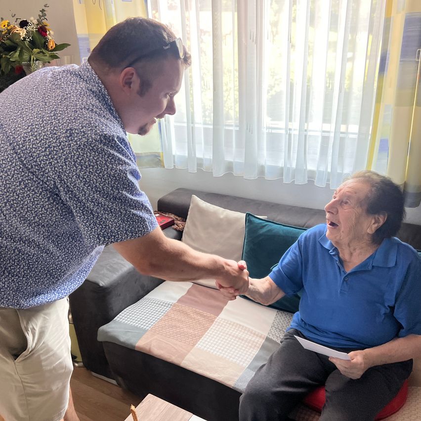 Zwei Personen schütteln sich in einem Raum die Hände. Ein älterer Mann hält Papier, während ein jüngerer Mann steht. Ein Sofa mit Kissen steht dahinter. Ein Blumenvase ist links.