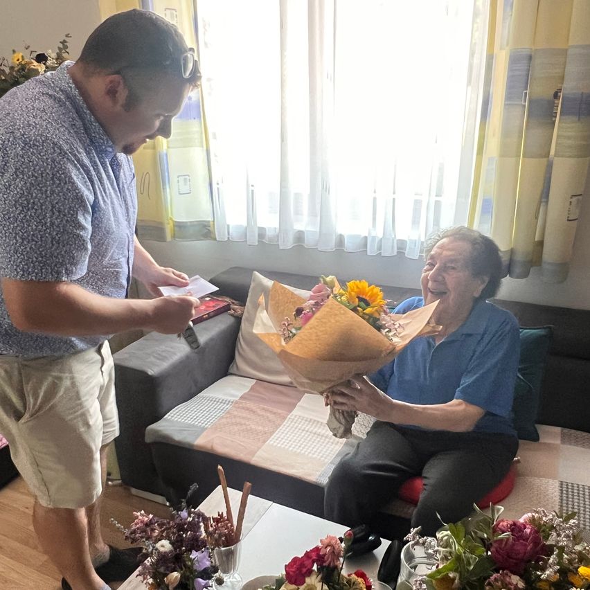 Eine ältere Frau hält einen Blumenstrauß und sitzt auf einer Couch, während ein Mann vor ihr steht. Der Mann hält ein Papier und einen Stift, wahrscheinlich um eine Karte zu unterschreiben. Die Couch steht in der Nähe eines Fensters mit weißen Vorhängen.