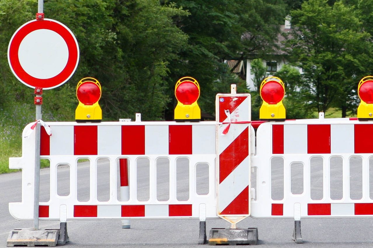 A red and white striped barrier with three red flashing lights on top is set up on the road, possibly indicating a road closure. Trees and a house are in the background.