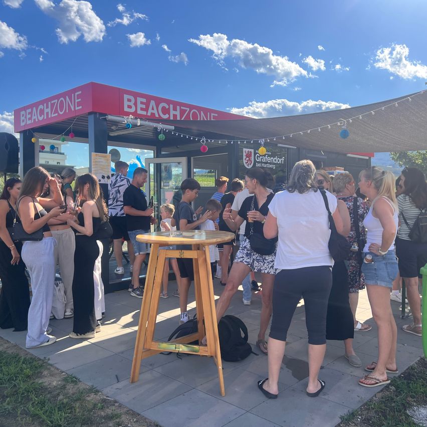 A group of people are gathered outside a Beachzone kiosk, some are standing and others are sitting on a stool. The kiosk has a red roof and a sign that reads Beachzone. There are also people standing near the entrance of the kiosk.