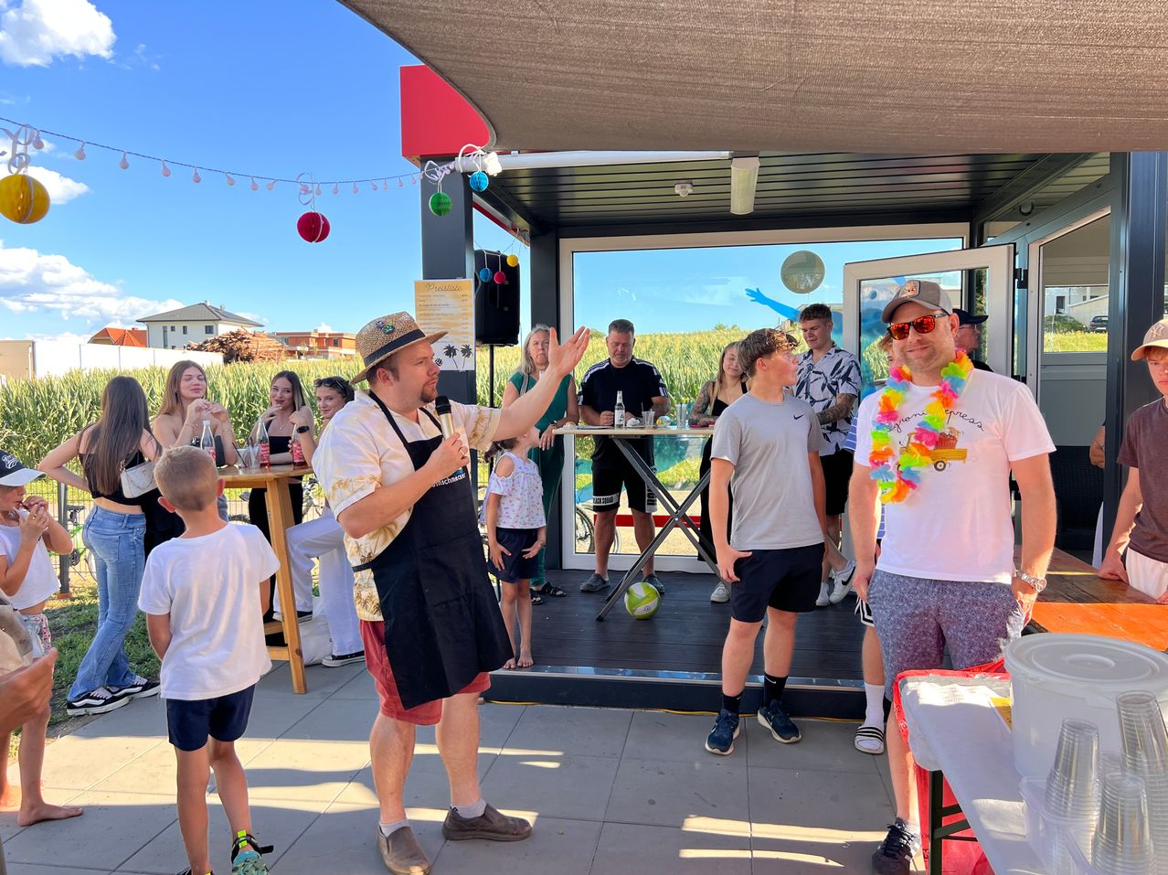 A group of people are gathered outdoors, with a man in a hat and apron speaking into a microphone. A boy in a white shirt stands next to him.