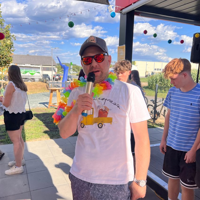 A man wearing a colorful lei and a white t-shirt with a cartoon truck on it speaks into a microphone. Behind him, two young boys and a woman stand. The scene is outdoors with a clear sky and some decorative lights.