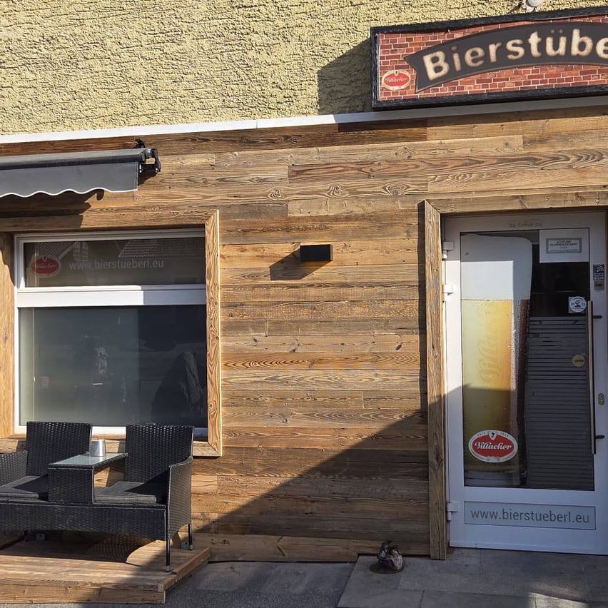 Ein Außensitzbereich mit zwei schwarzen Stühlen und einem Tisch vor einem Holzgebäude. Das Gebäude hat ein Glasfenster, eine Markise und ein Schild mit der Aufschrift 'Bierstuben'.