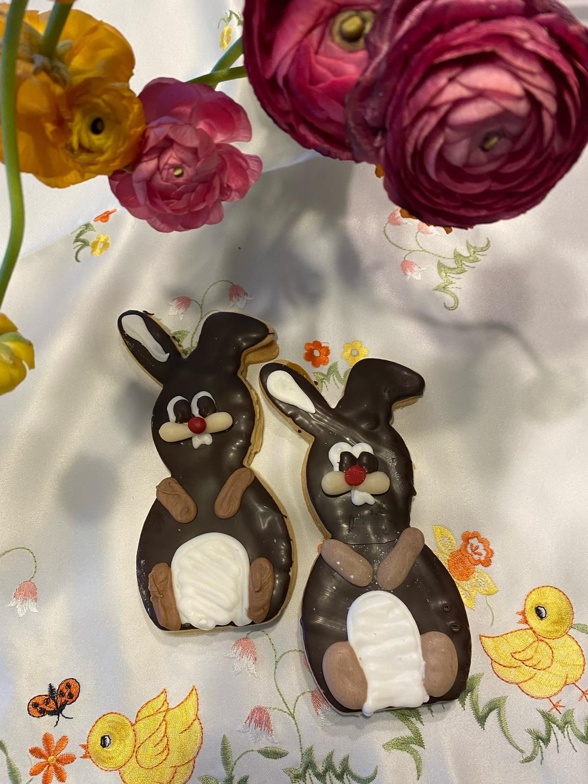 Zwei Schokoladenüberzogene Hasenkekse liegen auf einer blumengemusterten Tischdecke, mit Blumen im Hintergrund.