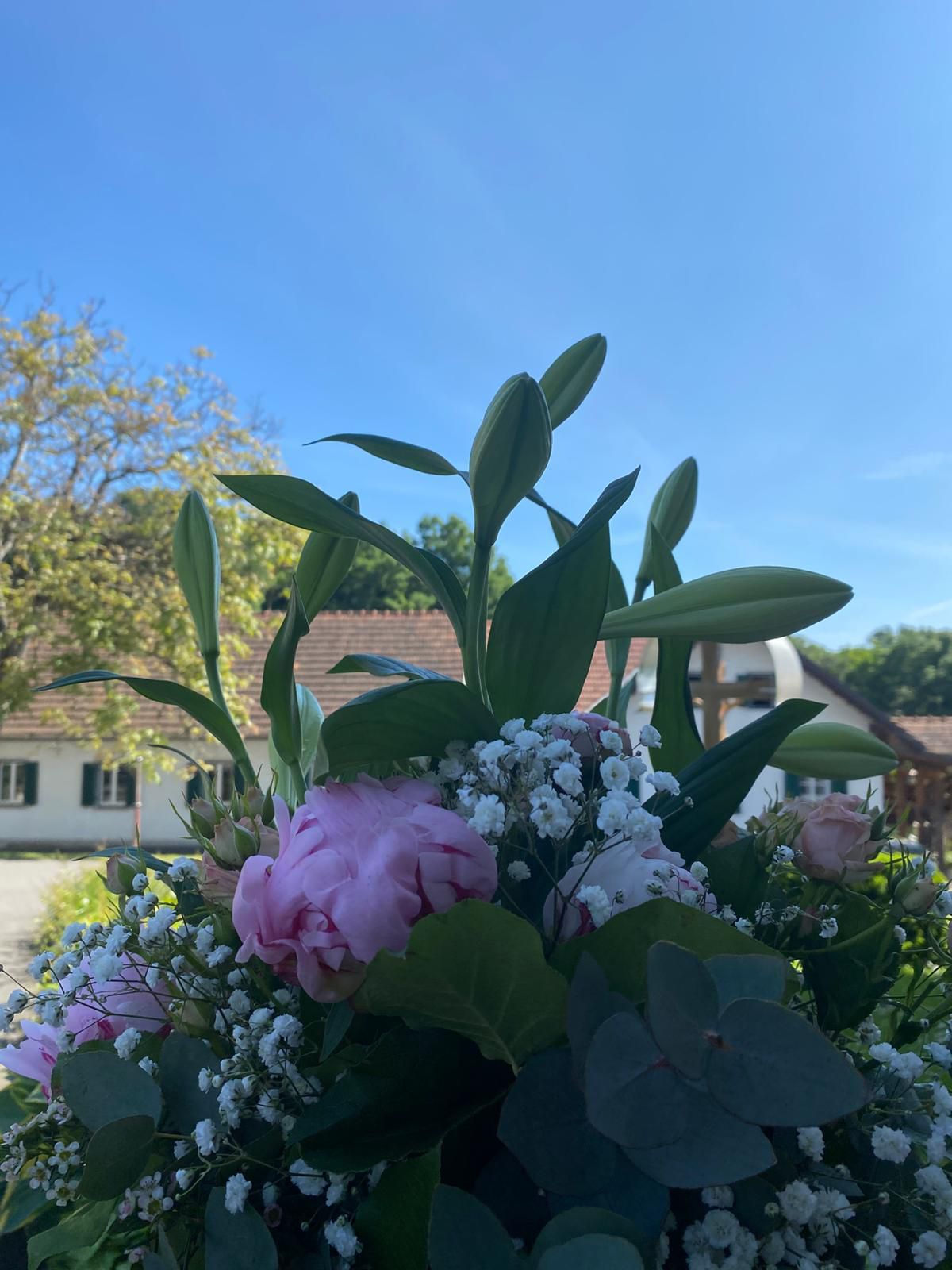 Ein Strauß aus rosa und weißen Blumen mit grünen Blättern steht vor einem Haus mit braunem Dach und weißen Wänden unter einem blauen Himmel.