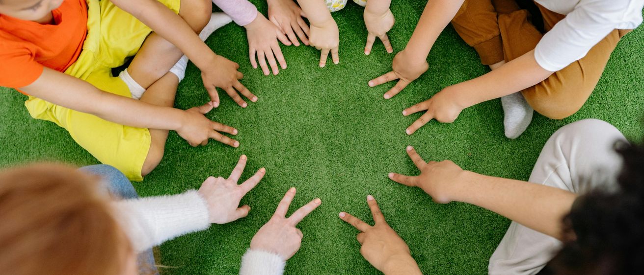 Eine Gruppe von Kindern im Kreis, Hände auf grünem Teppich, bilden ein V-Zeichen, tragen verschiedene Kleidung.