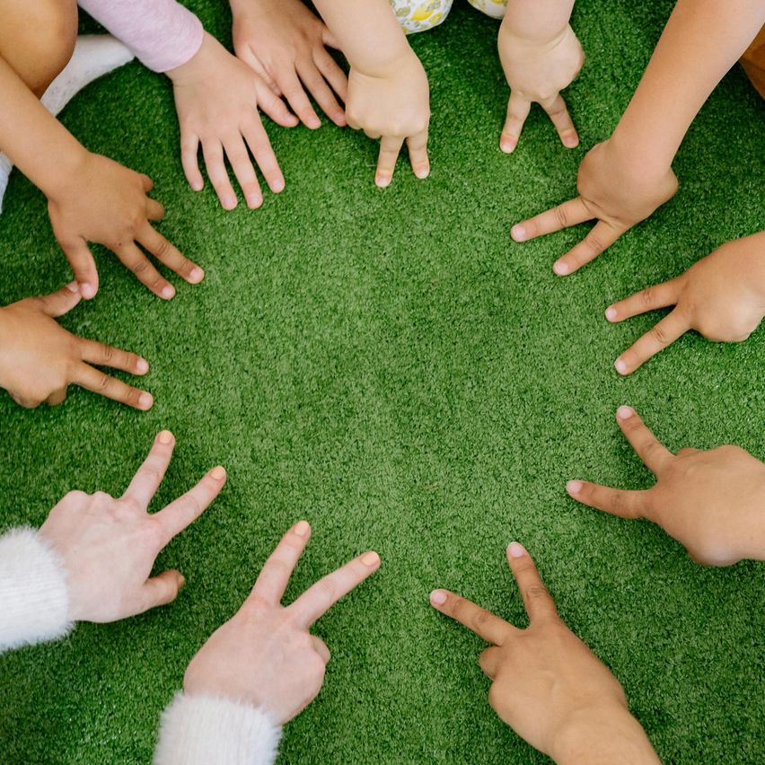 Eine Gruppe von Kindern im Kreis, Hände auf grünem Teppich, bilden ein V-Zeichen, tragen verschiedene Kleidung.