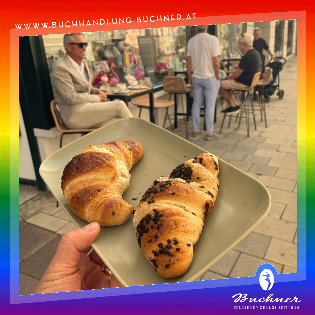 Eine Person hält einen Teller mit zwei schokoladenüberzogenen Croissants in einem Café. Hinter ihr sitzt ein Mann im Anzug an einem Tisch, und andere sitzen an hohen Stühlen. Das Café hat einen Ziegelboden und einen bunten Rand.