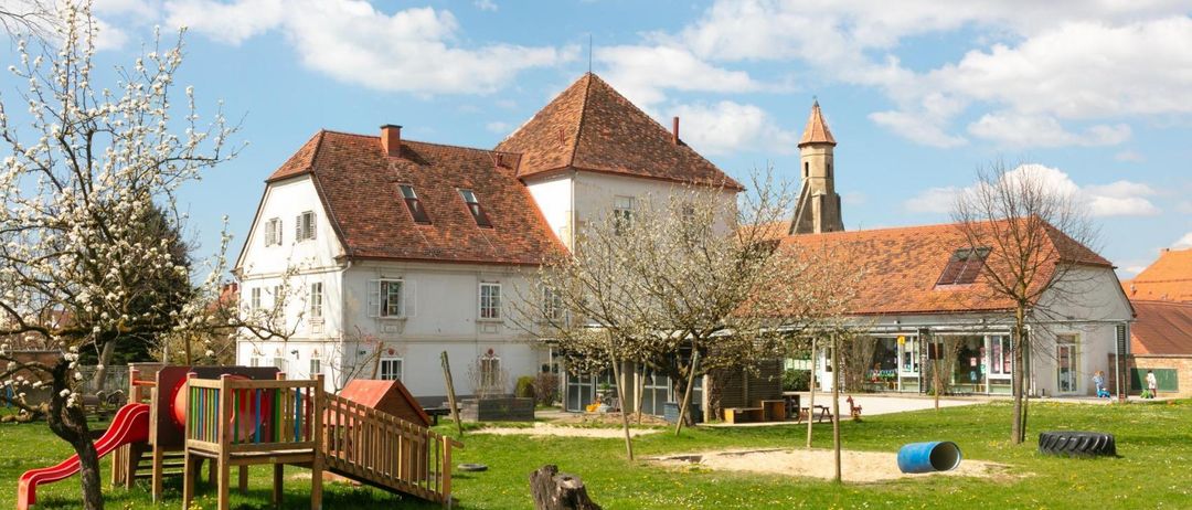 Ein großes weißes Gebäude mit braunen Dächern und Fenstern ist von einem Rasengebiet mit einer Holzrutsche und einem Baum umgeben.