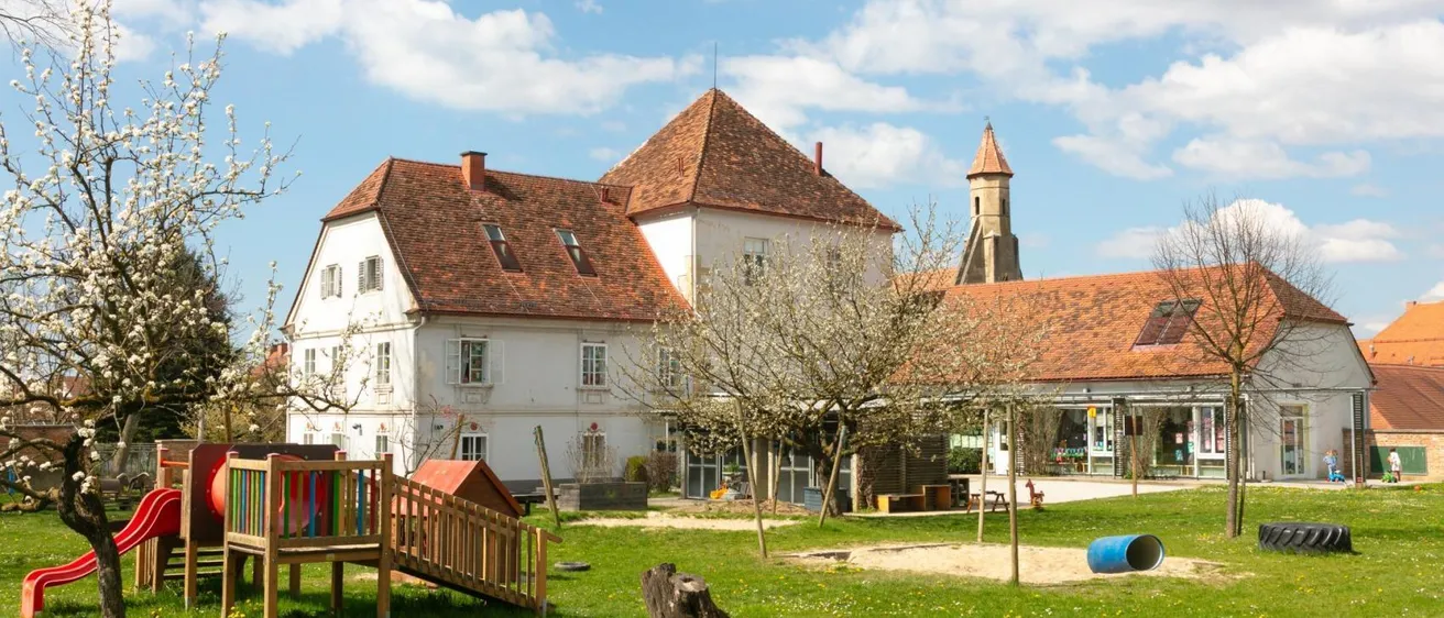 Ein großes weißes Gebäude mit braunen Dächern und Fenstern ist von einem Rasengebiet mit einer Holzrutsche und einem Baum umgeben.