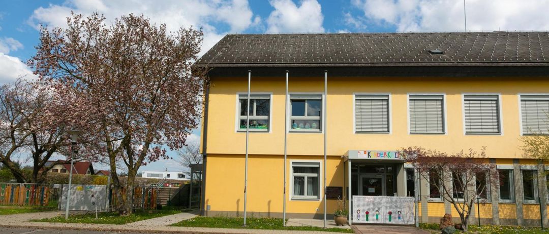 Ein leuchtend gelbes Gebäude mit einem grauen Dach hat drei Fenster und eine Tür. Die Fenster haben weiße Rahmen und Jalousien. Es gibt einen kleinen Garten mit einem Topfpflanzen und einem weißen Zaun.