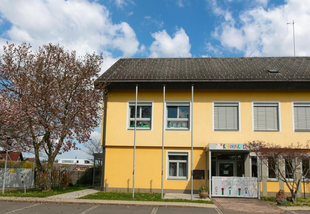Ein gelbes Gebäude mit schwarzem Dach und Glasfenstern. Es ist ein Kindergarten mit einem Spielplatz davor.