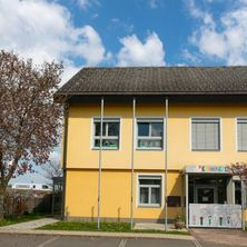Ein gelbes Gebäude mit schwarzem Dach und Glasfenstern. Es ist ein Kindergarten mit einem Spielplatz davor.