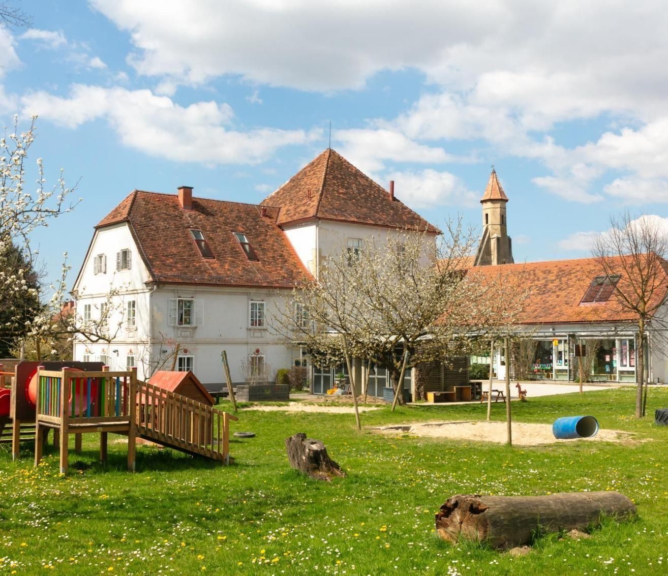 Ein großes weißes Gebäude mit braunen Dächern und Fenstern steht auf einem Rasen. In der Nähe gibt es ein hölzernes Spielhaus, einen Spielplatz und einen blauen Fass.