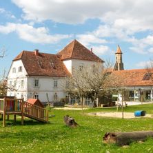 Ein großes weißes Gebäude mit braunen Dächern und Fenstern steht auf einem Rasen. In der Nähe gibt es ein hölzernes Spielhaus, einen Spielplatz und einen blauen Fass.