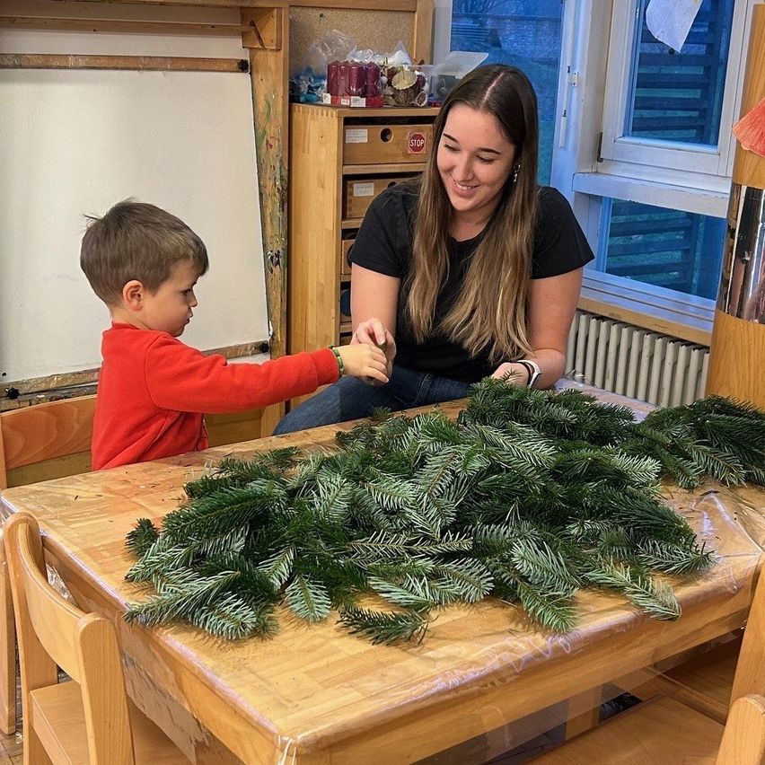 Ein junger Junge und eine Frau schmücken einen Tisch mit Tannengirlanden, lächeln und genießen die Aktivität. Der Raum hat Holzmöbel und eine Tafel.