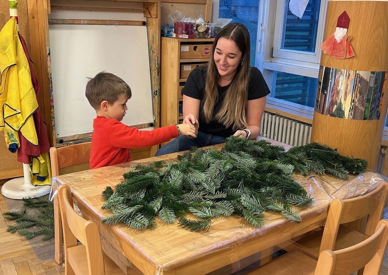 Ein junger Junge und eine Frau schmücken einen Tisch mit Tannengirlanden, lächeln und genießen die Aktivität. Der Raum hat Holzmöbel und eine Tafel.