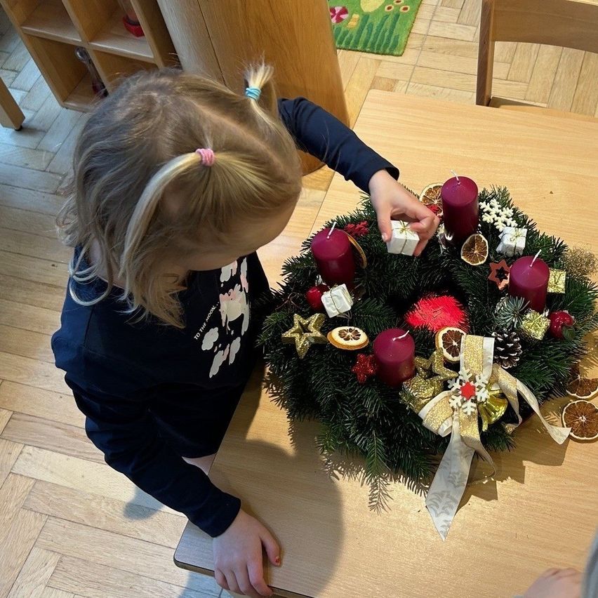 Ein kleines Mädchen schmückt einen Weihnachtskranz mit roten Kerzen und Ornamenten auf einem Holztisch.