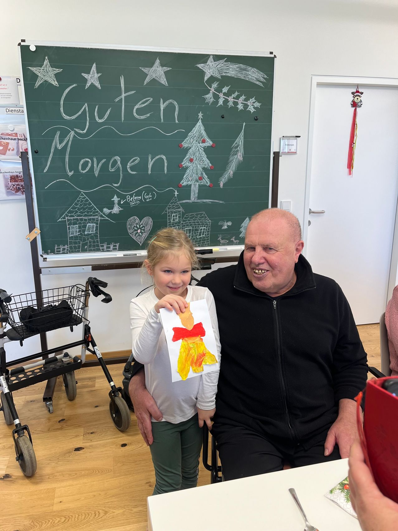 Ein Mann und ein junges Mädchen sitzen zusammen in einem Raum. Hinter ihnen zeigt eine Tafel 'Guten Morgen' in Kreide. Das Mädchen hält eine Zeichnung einer Figur mit einem roten Band.