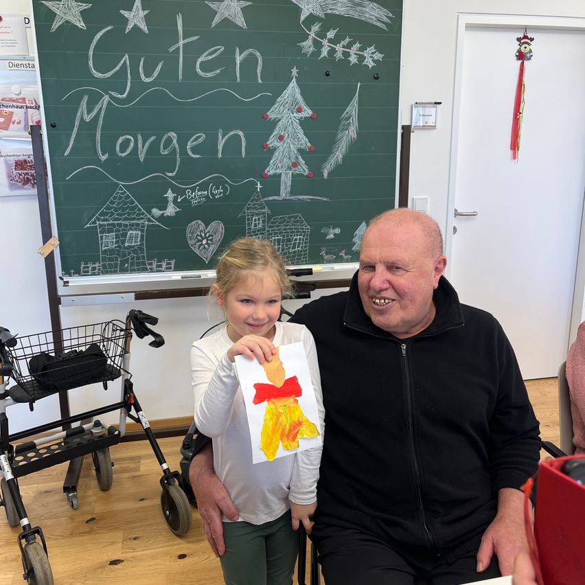 Ein Mann und ein junges Mädchen sitzen zusammen in einem Raum. Hinter ihnen zeigt eine Tafel 'Guten Morgen' in Kreide. Das Mädchen hält eine Zeichnung einer Figur mit einem roten Band.