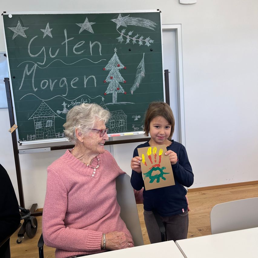 Eine ältere Frau mit einem rosa Pullover sitzt neben einem jungen Mädchen, das eine Papierhand mit einem gemalten Weihnachtsbaum in der Hand hält, beide lächeln in einem Raum mit einer Tafel, auf der 'Guten Morgen' steht.