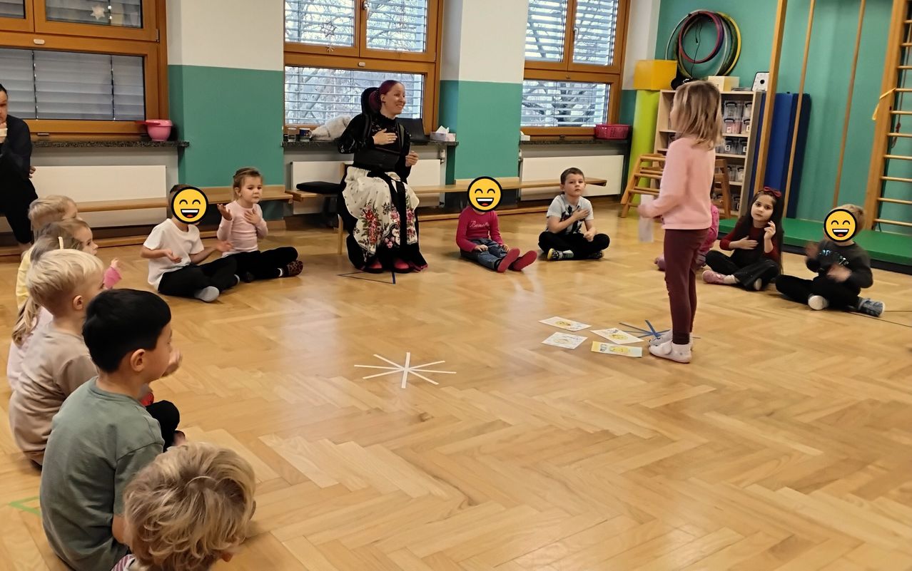 Eine Gruppe junger Kinder sitzt im Kreis auf dem Boden, mit einer Frau, die die Sitzung leitet. Die Kinder tragen Freizeitkleidung. Auf dem Boden sind Bücher und andere Gegenstände verstreut.