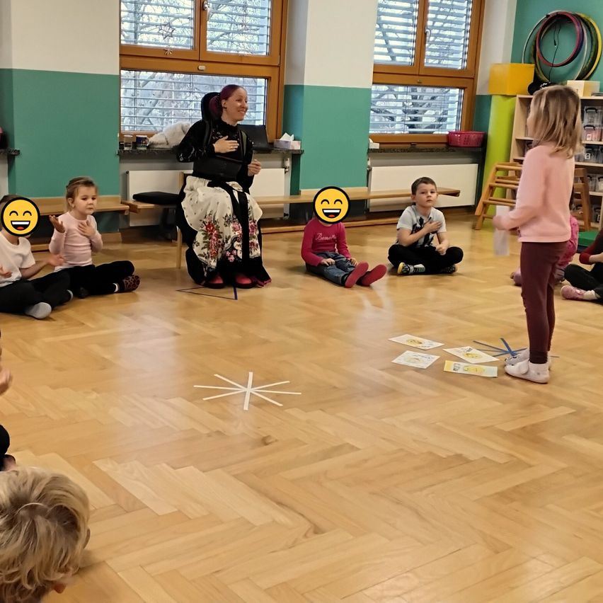 Eine Gruppe junger Kinder sitzt im Kreis auf dem Boden, mit einer Frau, die die Sitzung leitet. Die Kinder tragen Freizeitkleidung. Auf dem Boden sind Bücher und andere Gegenstände verstreut.
