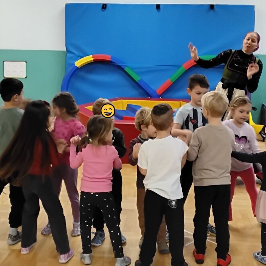 Eine Gruppe von Kindern tanzt in einer Turnhalle mit einer lächelnden Frau vor ihnen. Hinter ihnen befindet sich eine farbenfrohe Wand.