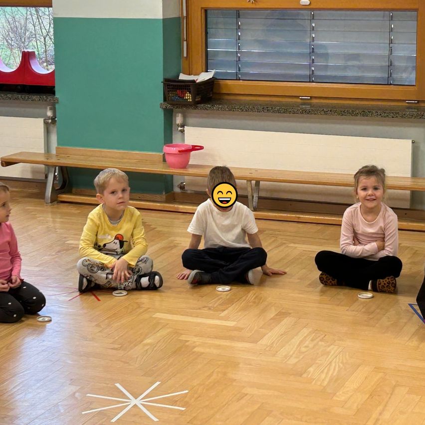 Vier Kinder sitzen auf dem Boden in einem Raum mit einer Holzbank und Fenstern. Ein Kind hat ein lächelndes Gesicht.