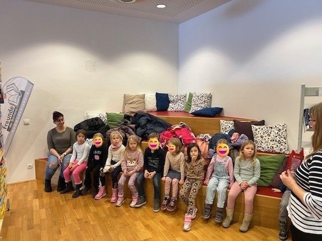 Eine Gruppe von Kindern sitzt auf einer Bank mit Kissen und Jacken. Zwei Frauen stehen in der Nähe. Die Kinder tragen Stiefel und Schals.