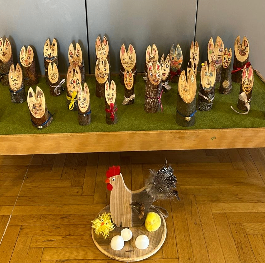 Ein Holztisch mit einem grünen Teppich hat verschiedene Holzskulpturen von Kaninchen darauf. Unten auf dem Tisch befindet sich ein kreisförmiger Holzteller mit einem Hahn und einigen Eiern.