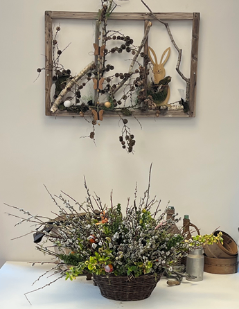 Ein Korb mit Blumen steht auf einem Tisch. Ein an der Wand montierter Holzrahmen mit einem Hasendesign, Tannenzapfen und Zweigen befindet sich über dem Tisch. Ein silberner Kanister und ein Holzkorb befinden sich neben den Blumen.