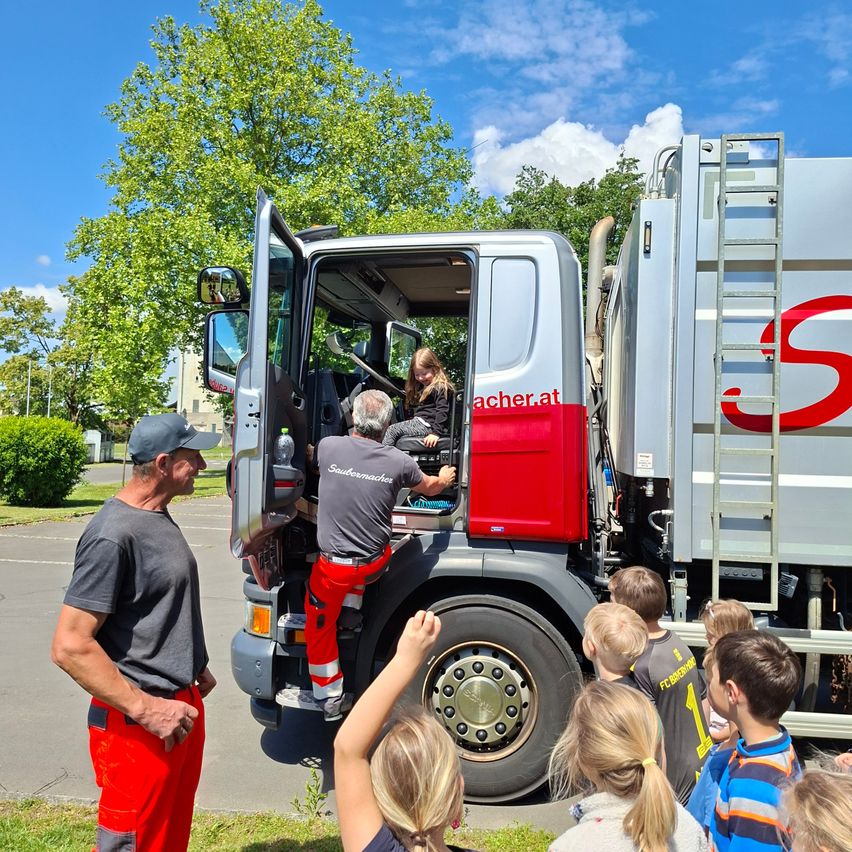 Eine Gruppe von Kindern steht um einen Feuerwehrwagen. Ein Feuerwehrmann hilft einem Kind, in den Wagen zu steigen.