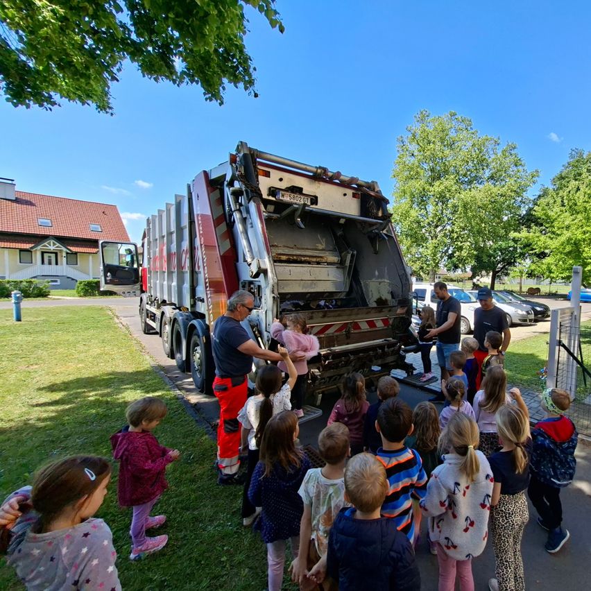 Eine Gruppe von Kindern beobachtet, wie ein Müllwagen geleert wird. Ein Mann zeigt ihnen, wie er funktioniert.