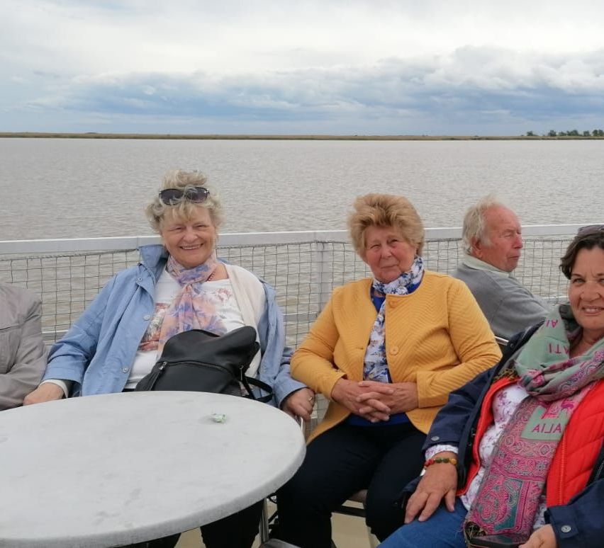Vier ältere Menschen sitzen auf einer Terrasse am Wasser. Eine Frau in Blau lächelt neben einer in Gelb. Dahinter schauen zwei Männer zum Wasser.