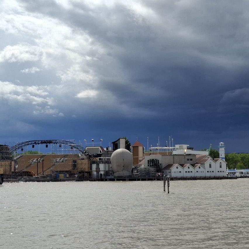 Ein Wasserfrontbereich mit bewölktem Himmel. Es gibt Gebäude und Strukturen mit einem großen Bogen und einer Kuppel. Ein Pfosten steht im Wasser.