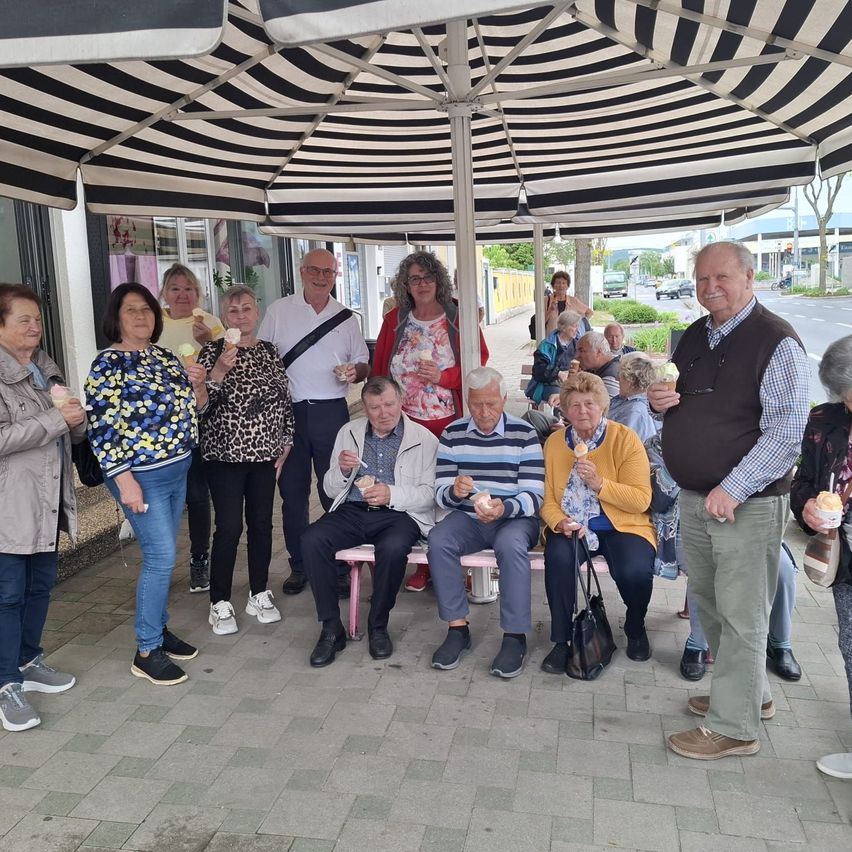 Eine Gruppe von Menschen, sowohl Männer als auch Frauen, sitzen auf einer Bank unter einem Sonnenschirm und halten Eisbecher in der Hand.