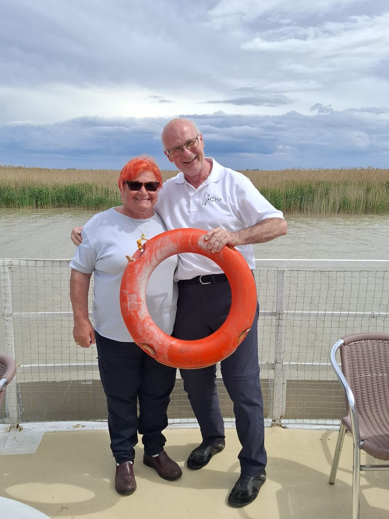 Ein Paar steht auf einem Deck mit einer Brüstung und lächelt. Ein Mann hält einen roten Rettungsring. Hinter ihnen ist Wasser und ein Grasbereich unter einem bewölkten Himmel.