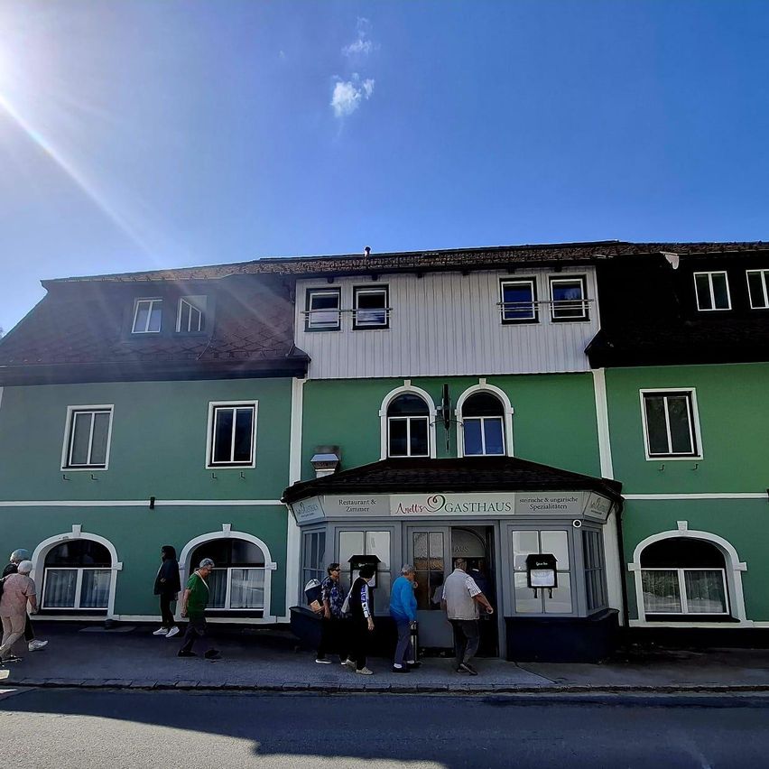 Ein sonniger Tag mit Menschen, die an einem grünen Gebäude mit Restaurant-Schild und mehreren Rundbogenfenstern vorbeigehen.