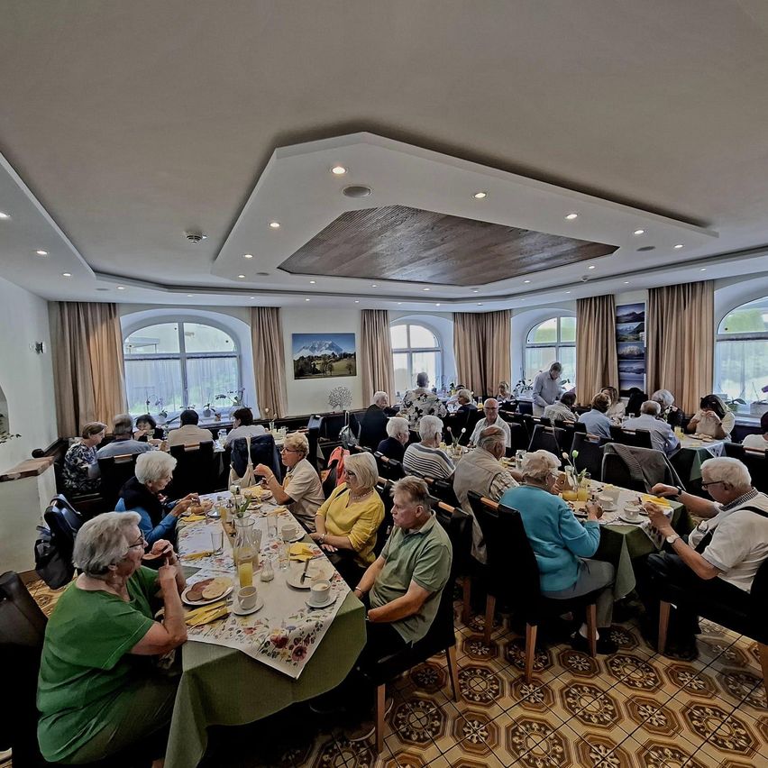 Eine große Gruppe älterer Menschen diniert gemeinsam in einem Restaurant. Tische sind mit verschiedenen Speisen besetzt, und Menschen sitzen darum herum. Ein Gemälde hängt an der Wand, und das Restaurant hat hohe Decken mit Beleuchtung.