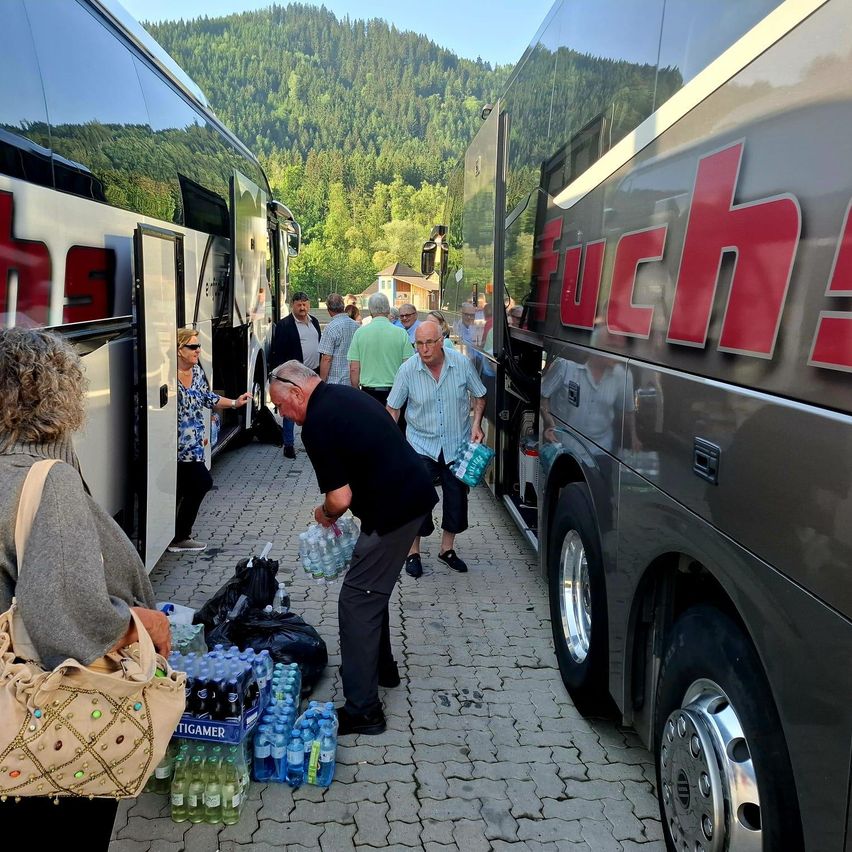 Mehrere Personen laden Gegenstände von einem Bus aus. Ein Mann beugt sich, um Wasserflaschen aufzuheben. Eine Frau trägt eine Handtasche.