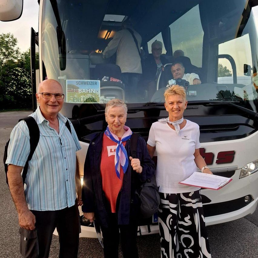 Drei Personen stehen vor einem Bus. Ein Mann links trägt eine Brille und einen Rucksack. Eine Frau in der Mitte hat ein Tuch und eine Tasche. Eine Frau rechts hält ein Papier. Ein Mann im Bus trägt ein weißes Hemd.
