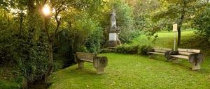 Ein ruhiger Park mit einem kleinen Bach, zwei Holzbänken und einer Statue, umgeben von üppigem Grün unter einem hellen, sonnigen Himmel.