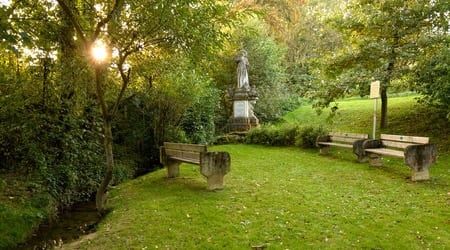 Ein ruhiger Park mit einem kleinen Bach, zwei Holzbänken und einer Statue, umgeben von üppigem Grün unter einem hellen, sonnigen Himmel.