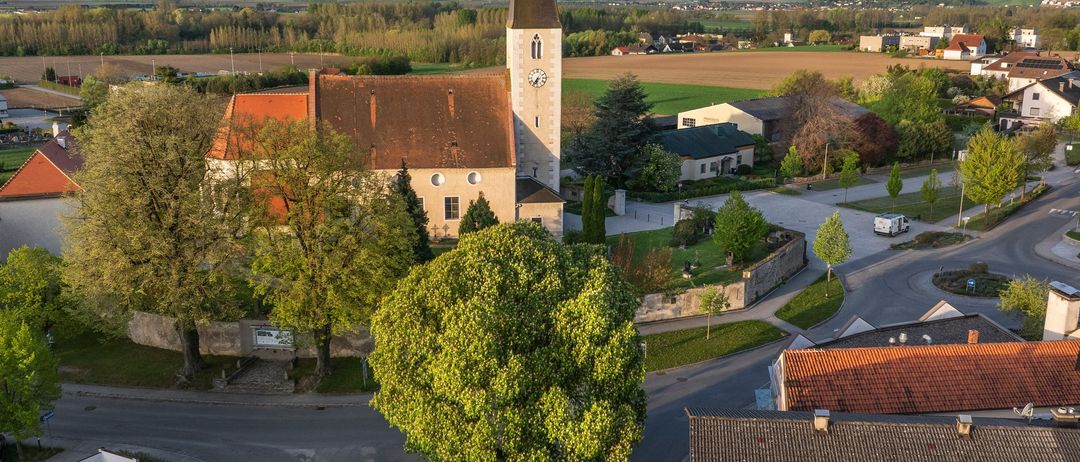 Luftaufnahme einer Kirche mit einem hohen Turm und einer Uhr, umgeben von Bäumen und Gebäuden. Die Kirche befindet sich in einer ländlichen Gegend mit einem weiten Feld und Bergen in der Ferne.
