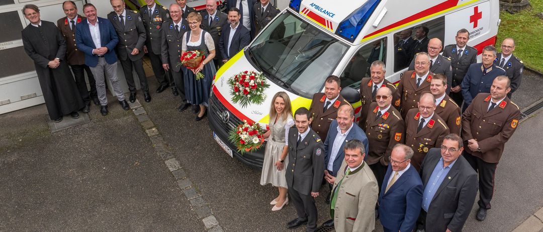 Eine Gruppe von Menschen, darunter eine Frau in einem weißen Kleid, die Blumen hält, posiert für ein Foto vor einem Krankenwagen.