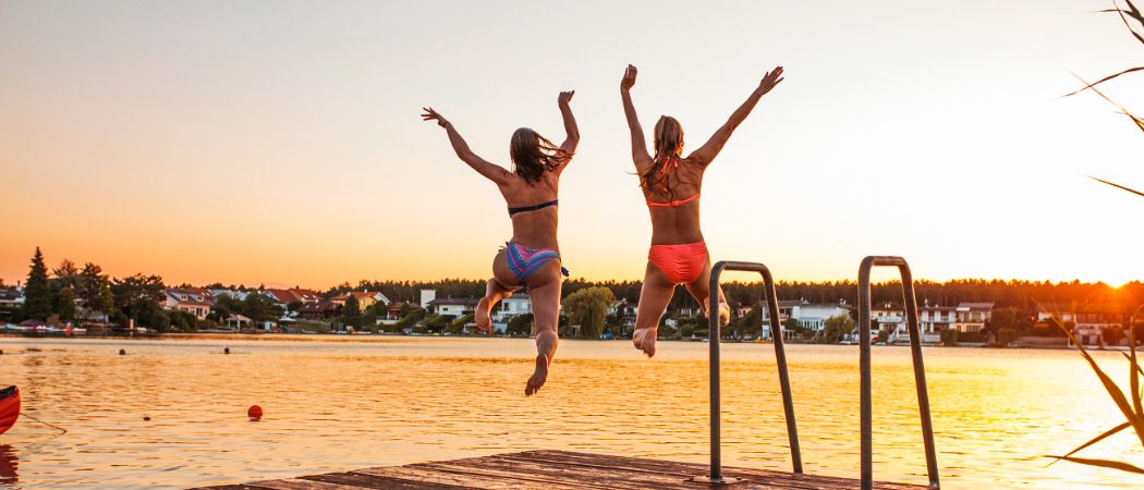 Zwei Frauen springen bei Sonnenuntergang von einem Steg in einen See, mit Häusern und Bäumen im Hintergrund.