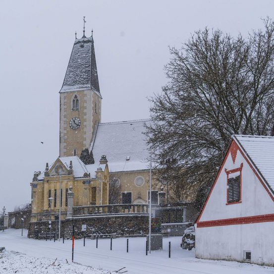 Eine Kirche mit einem Glockenturm und einem kleinen Haus, das mit Schnee bedeckt ist. Die Kirche hat ein Spitzdach und einen Balkon.