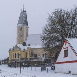 Eine Kirche mit einem Glockenturm und einem kleinen Haus, das mit Schnee bedeckt ist. Die Kirche hat ein Spitzdach und einen Balkon.